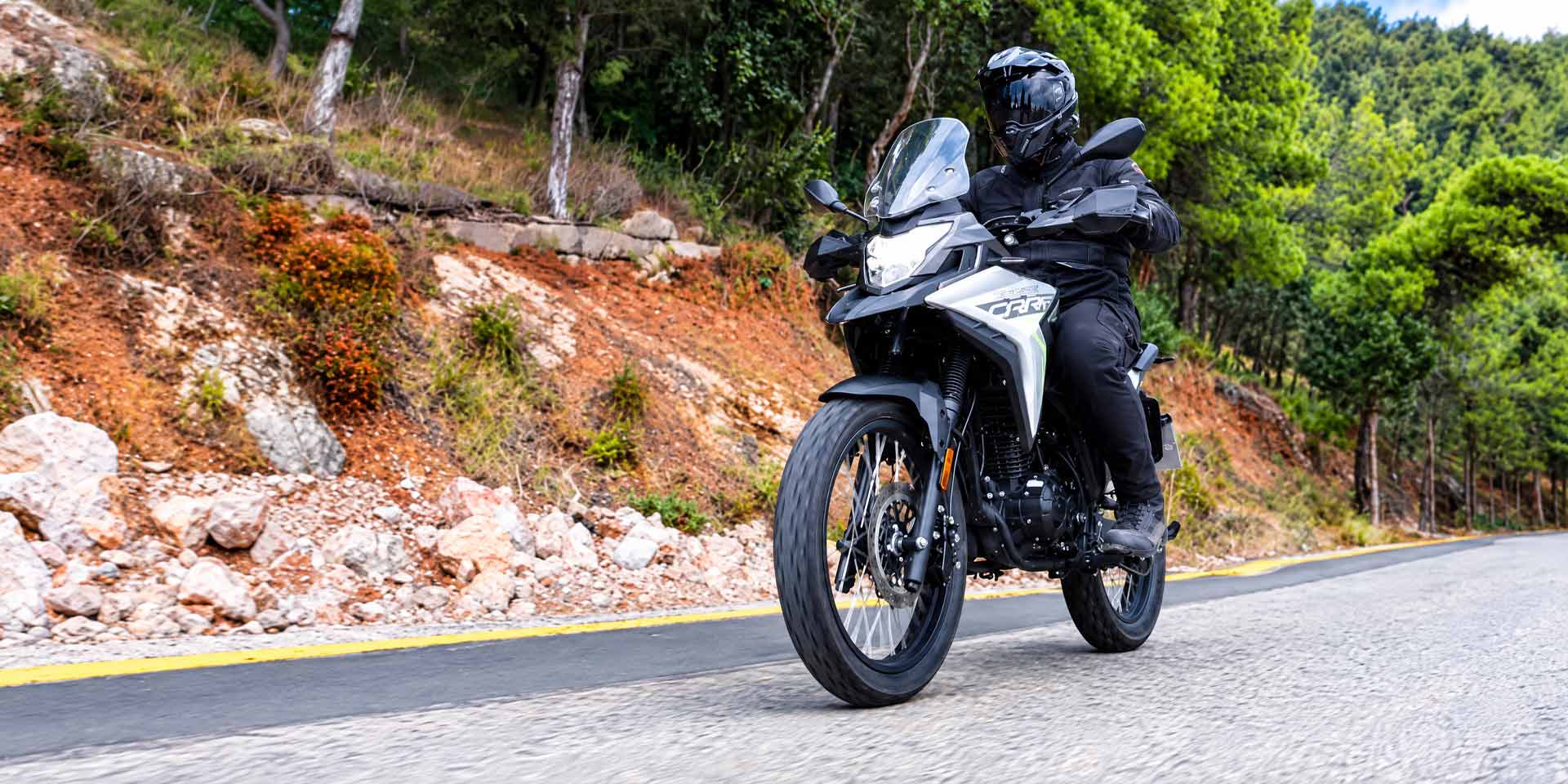 Αναβάτης με μαύρο εξοπλισμό οδηγεί το Cyclone Carrera 125 σε δρόμο με πεύκα και κοκκινωπό πρανές στο φόντο